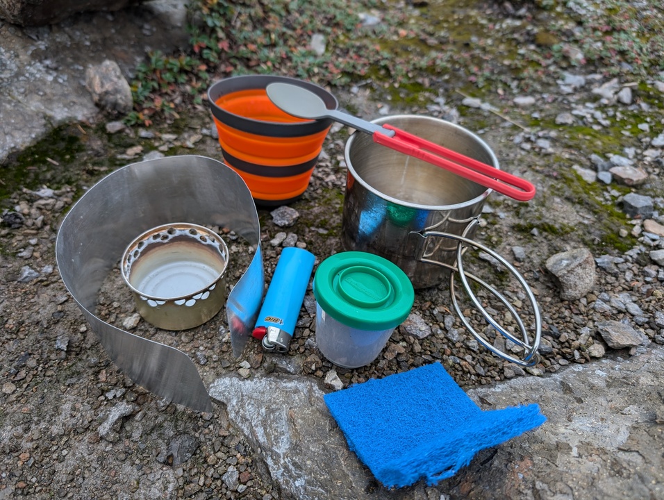 Cook set components (stove, wind screen, lighter, cook pot, cup, spoon, sponge, and coffee) laid out on rocks