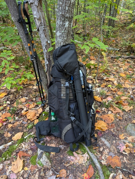 Backpacking pack with fishing rods on the outside and trekking poles leaning against a tree